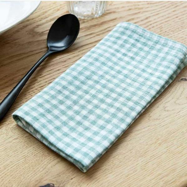 A set of four rosemary green and white gingham linen napkins styled on an oak table with black cutlery.