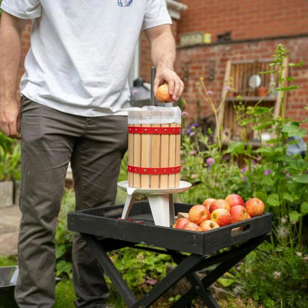 KuKoo fruit crusher with wooden barrel and red accents, a person adding apples, surrounded by a garden and fresh apples on a tray