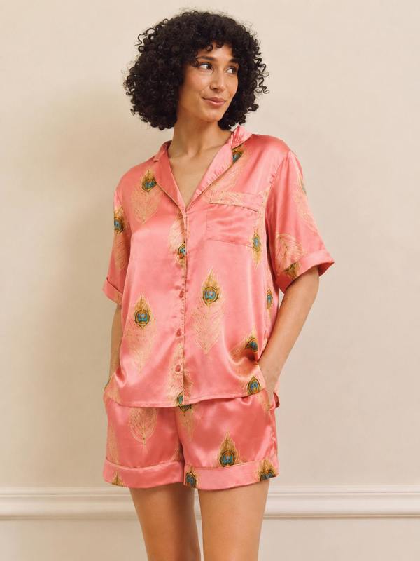 A woman wearing a two-piece satin pyjama set in pink with a gold and green peacock feather pattern, standing against a plain beige wall.