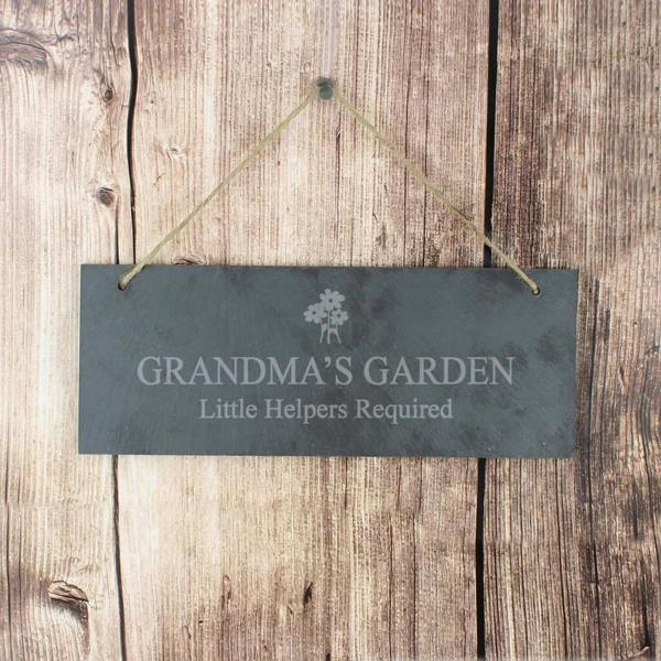 A rectangular hand-cut grey slate plaque engraved with a flower motif and "Grandma's Garden" hanging by a jute string on a wood panel background.