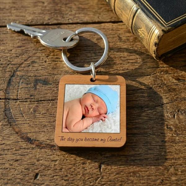 A square cherry wood keyring resting on a rustic old wooden table next to a vintage book and a silver key. The keyring features a photo of a sleeping newborn baby in a light blue hat, with the custom text 'The day you became my Auntie!' engraved underneath.