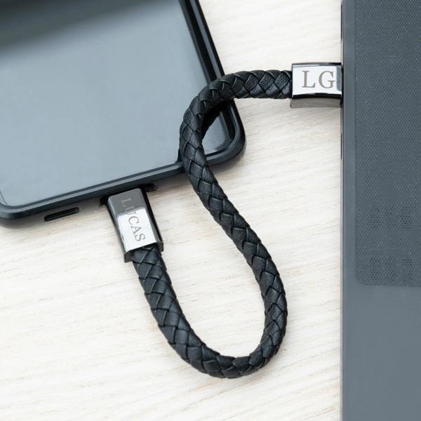 An unclasped braided black leather men's charger bracelet serving as a charging cord plugged between a black smartphone and a laptop terminal.
