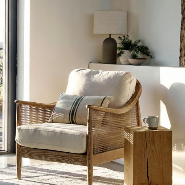 A Mid-Century Modern rattan armchair with a light oak wood frame and off-white linen cushions, styled in a bright sunlit corner next to a rustic wooden side table and a ceramic lamp.