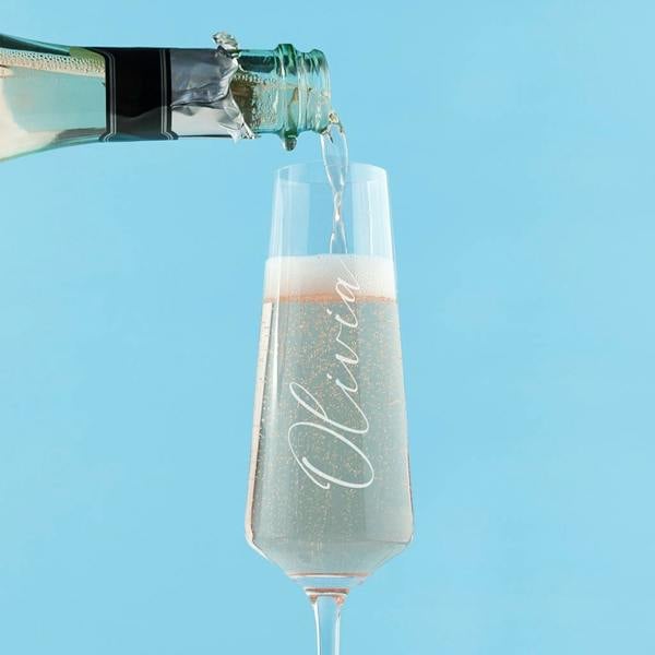 An elegant clear glass champagne flute custom engraved with a name in a vertical upward script font, sitting on a clean surface.