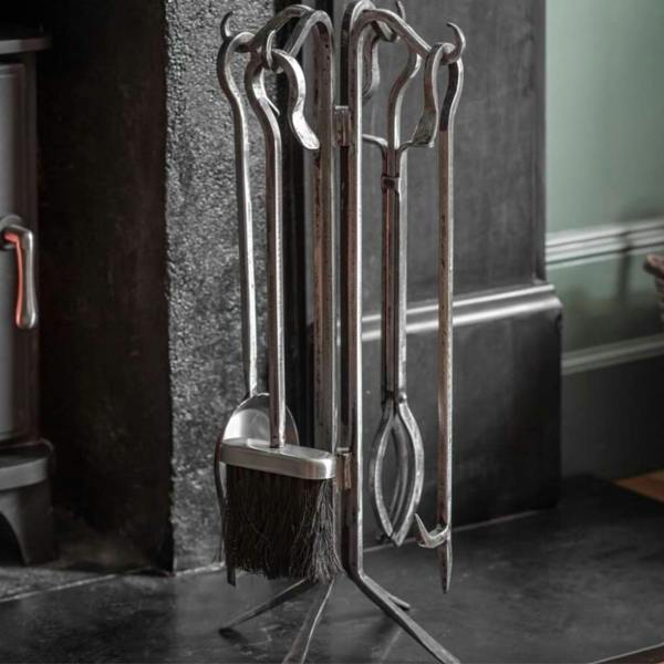 A freestanding antique pewter cast iron fireside companion tool set featuring a matching brush, shovel, tongs, and poker hanging on a central metal rack, positioned on a dark slate hearth next to a log-burning stove.