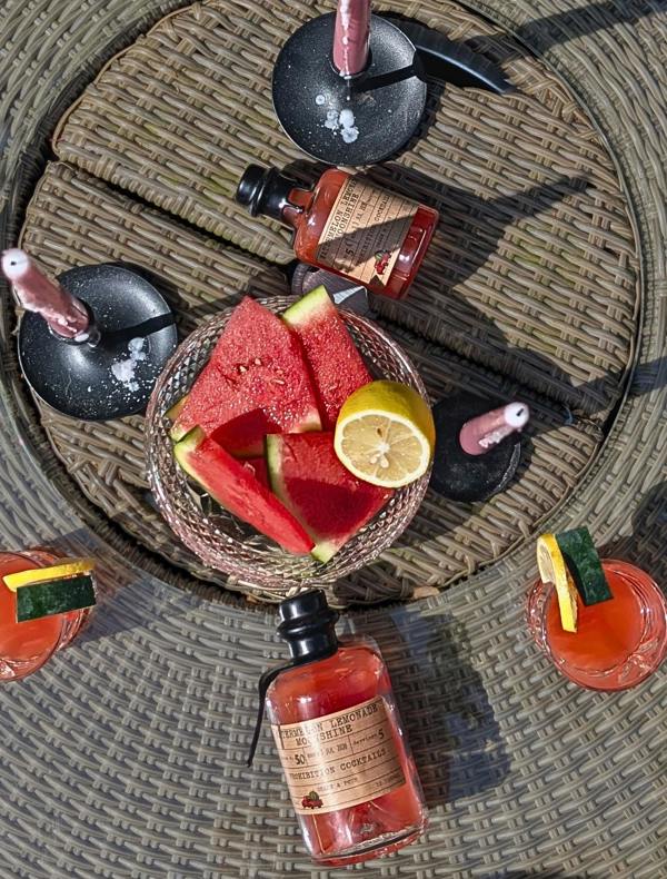 A styled overhead flat-lay of a cocktail-making setup on a woven round table. A glass bowl holds watermelon slices and a lemon wedge, surrounded by several bottles of pink-red liquids and garnished drinks with cucumber and lemon slices.