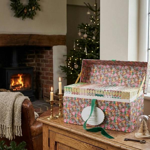 The open Liberty Festive Gifts Advent Calendar chest displayed on a rustic wooden console, surrounded by lit candles, fir branches, a glowing fireplace, and a decorated Christmas tree.