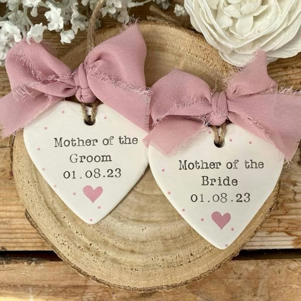 Two hand-stamped white ceramic heart decorations reading Mother of the Groom and Mother of the Bride with a wedding date, topped with dusty pink frayed ribbons on a wooden log slice.