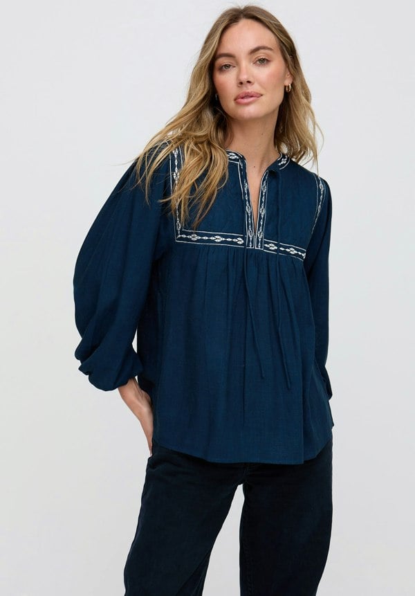 Woman wearing a navy blue blouse with white embroidery on a light gray background