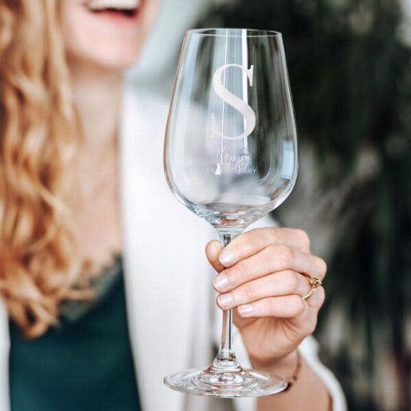 A woman laughing while holding up a personalised monogram wine glass engraved with the initial S.