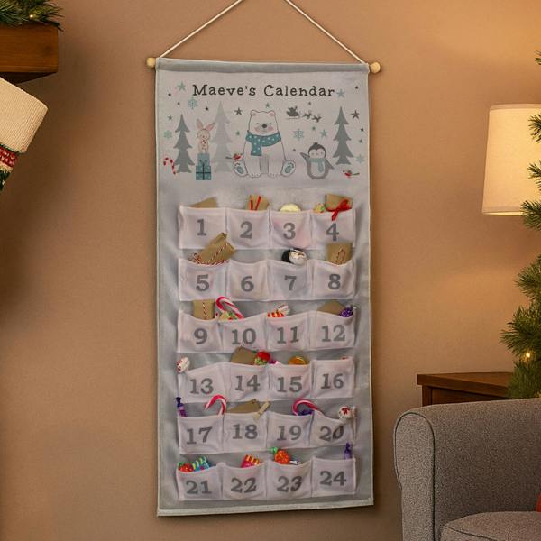 A fabric advent calendar hanging on a warm beige wall next to a grey armchair, featuring a polar bear illustration at the top with the text Maeve's Calendar, and 24 numbered white pockets filled with treats and small wrapped surprises.