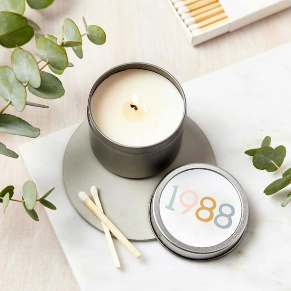 A silver metal tin candle on a grey stone coaster with a lid personalised with the year '1988' in pastel colours, accompanied by two matches and eucalyptus sprigs.