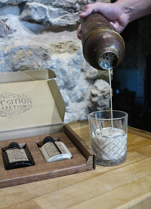 A hand pours a drink from a metal shaker into a clear textured glass on a wooden table, with a branded box and two small bottles in the background against a stone wall.