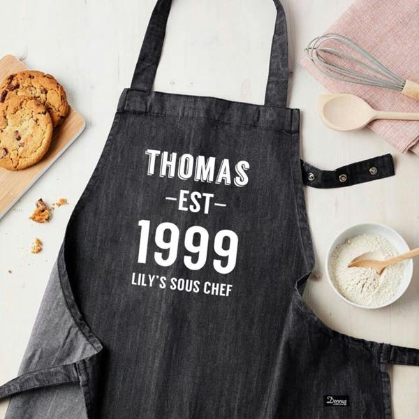 A dark grey denim apron reading Thomas Est 1999 Lily's Sous Chef laid flat next to a bowl of flour and chocolate chip cookies.