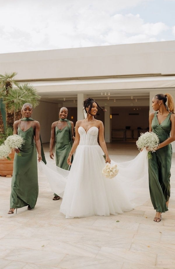 Bride in a white dress with three bridesmaids in green dresses, all holding bouquets outdoors.