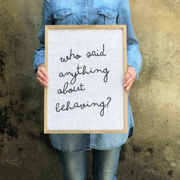 A woman holding a large solid oak framed print made of white cotton fabric with the lowercase handwritten-style text "who said anything about behaving?"