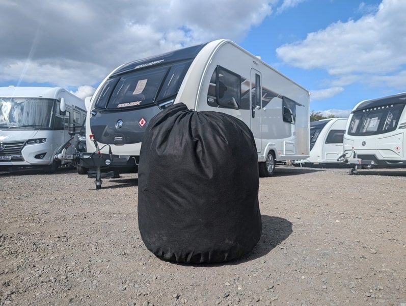 An image of the storage bag of the Breathable Waterproof 4Ply Caravan Cover with Free Hitch Cover Black from OLPRO pictured in front of a caravan in storage.