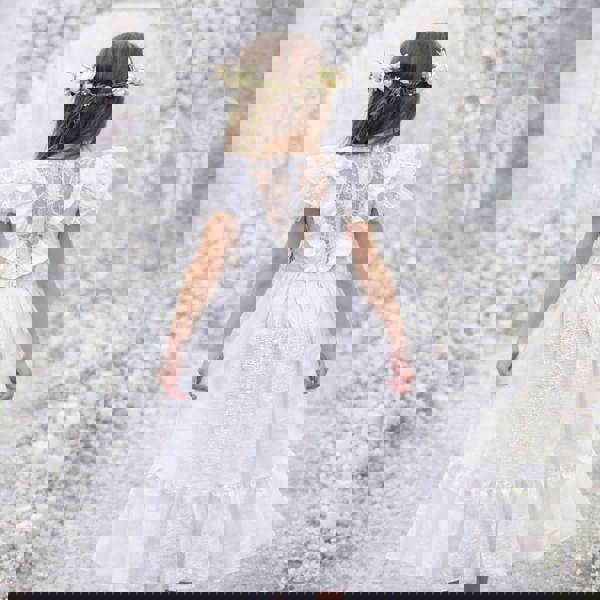 Young girl in a white dress with lace details standing against a textured white background.