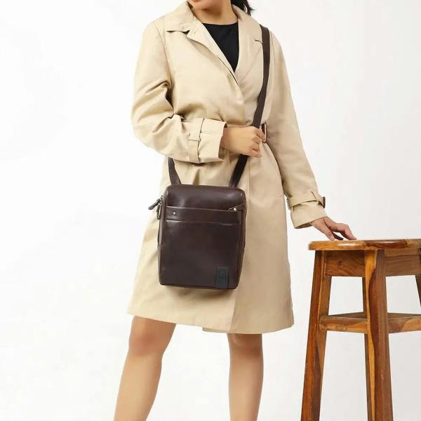 Person wearing a beige trench coat holding a brown leather messenger 
 bag on a white background
