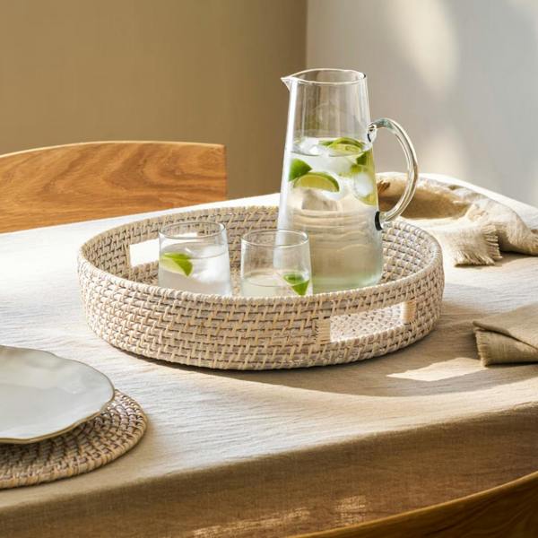 Round whitewash rattan tray styled with a water pitcher and glasses on a linen tablecloth.
