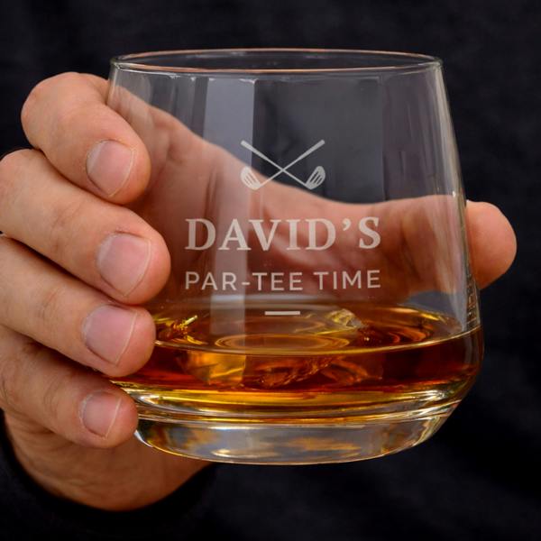 A hand holding a personalised glass whiskey tumbler filled with amber liquid and ice, featuring an engraved golf club logo and text that reads "David's Par-Tee Time."