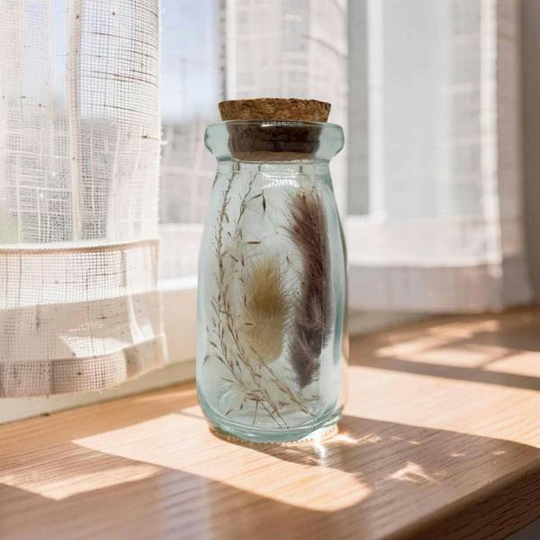 Glass jar with cork lid containing dried plants on a wooden surface with curtains in the background