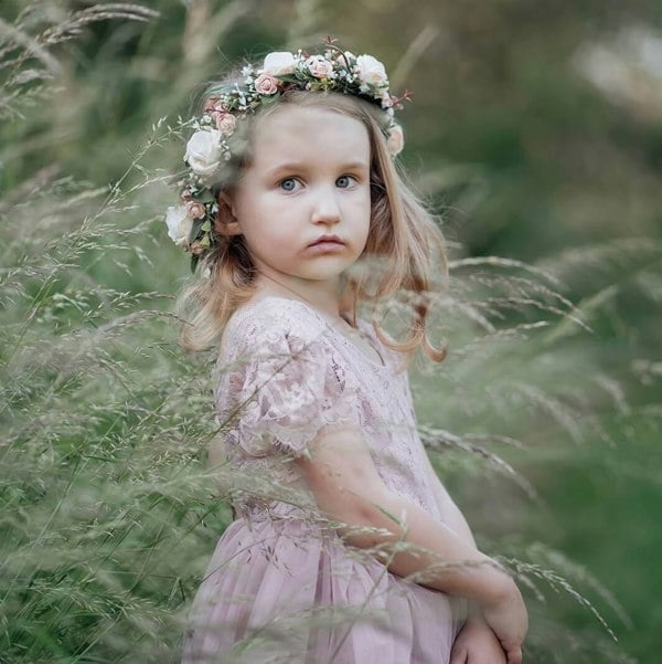 Young girl in a pink dress with a floral headband standing in tall grass.