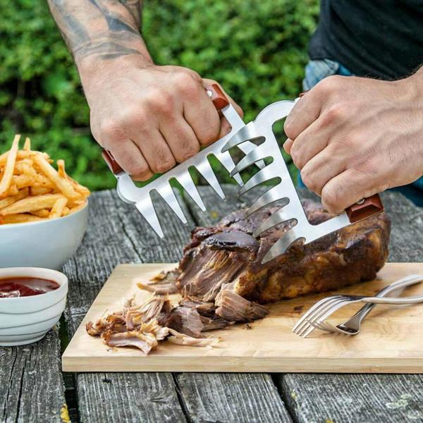 A close-up of a person's hands using two metal meat claws with wooden handles to shred a large piece of cooked pulled pork on a wooden cutting board outdoors next to a bowl of chips and sauce.