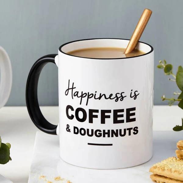 A white ceramic mug with a black handle and rim sitting on a marble coaster. The text reads 'Happiness is COFFEE & DOUGUNUTS'. Custard cream biscuits are on the side.