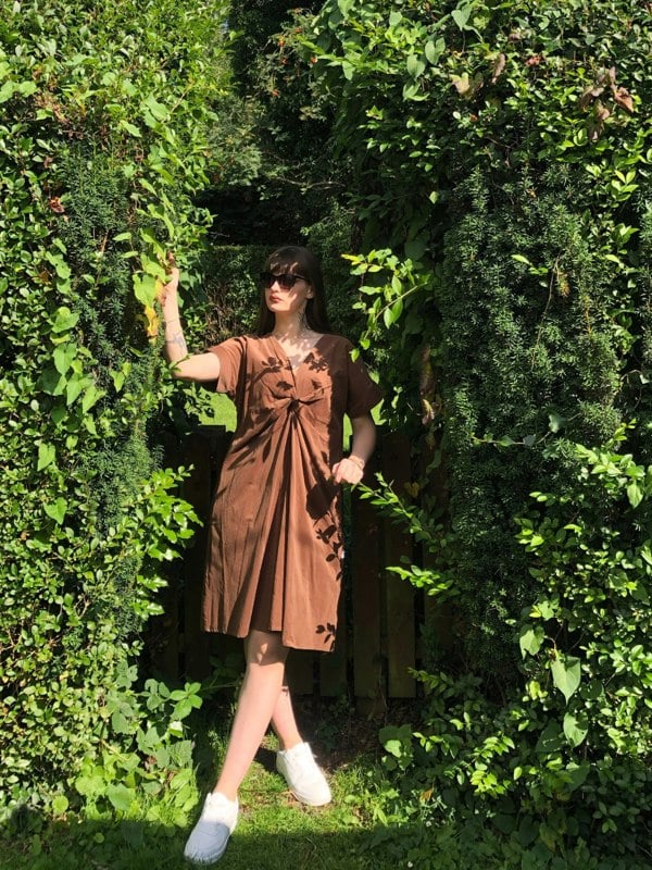 Pictured is the Riverbank Dress With a Knot in Brown Corduroy. Our model is standing against a fence in the woodlands. It is a Midi length dress, worn with white trainers