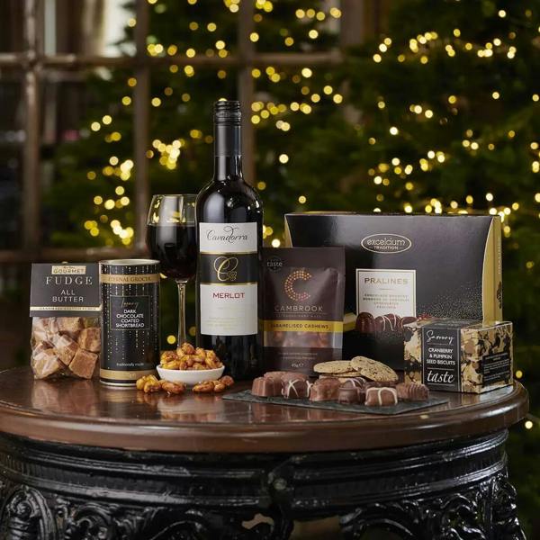 The Celebration Christmas Gift Box displayed on a dark wooden table with a bottle of Cavadorra Merlot, wine glass, chocolates, fudge, and nuts in front of a glowing Christmas tree