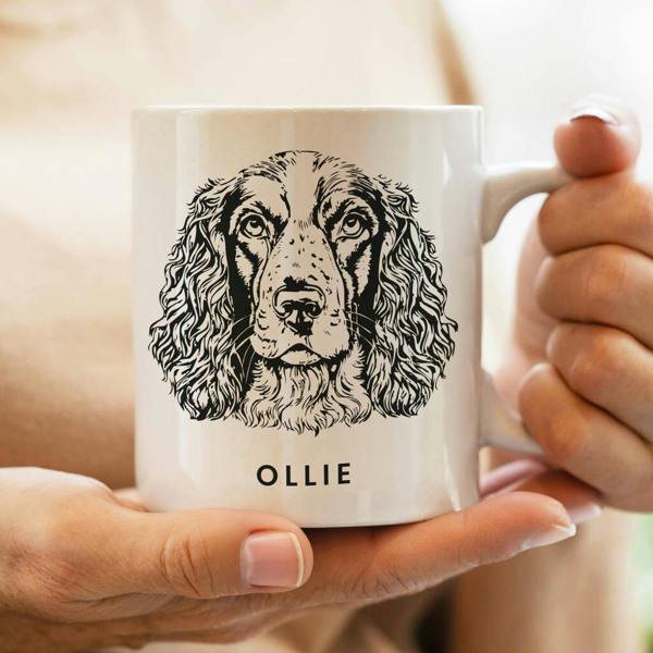 A person holding a white ceramic mug featuring a detailed black ink illustration of a Cocker Spaniel or Springer Spaniel with the name OLLIE printed underneath.