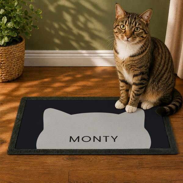 A tabby cat sitting beside a black personalised cat bowl mat featuring a grey cat silhouette graphic and the name MONTY.