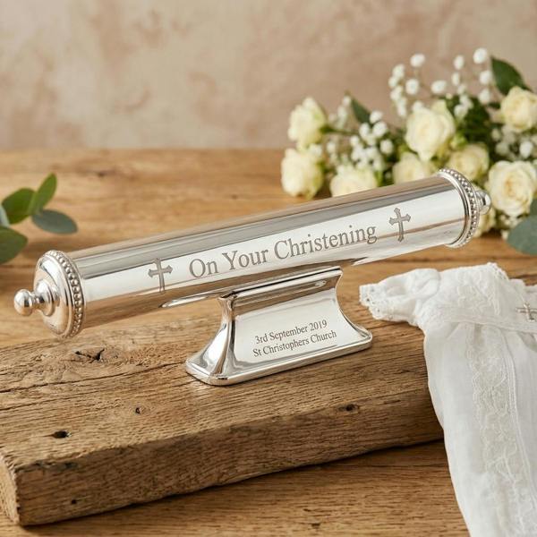 A personalised silver-plated christening certificate holder engraved with a custom date and church name, resting on an old farmhouse wooden surface with white flowers and a baby gown in the background.