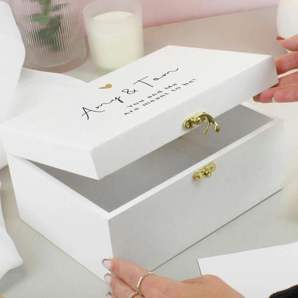 White wooden keepsake memory box with gold latches and a small gold heart graphic, personalised on the lid with the names 'Amy & Tom' and the text 'You and me Are meant to be!', being opened by a person's hands.