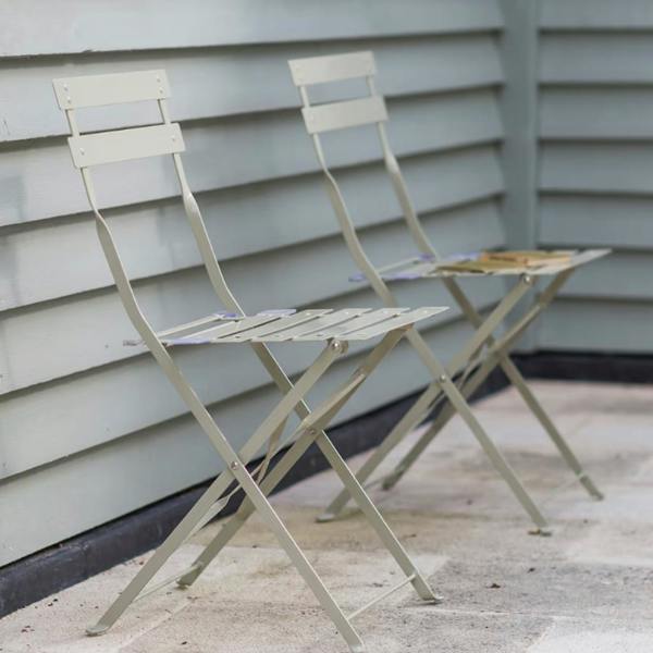 A pair of clay-coloured metal folding bistro chairs styled outdoors against pale green wooden cladding.