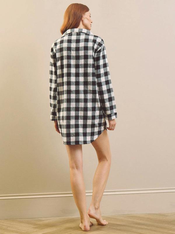 A woman with red hair is shown from behind wearing a black and white gingham check plaid long-sleeve nightshirt dress, standing barefoot on a light wood floor against a beige wall.