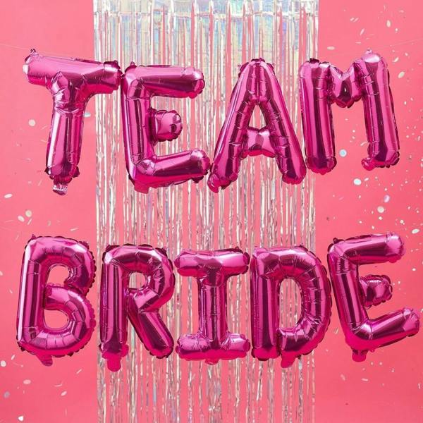 Hot pink foil letter balloons spelling out Team Bride arranged against a matching pink backdrop with iridescent silver fringe tinsel and scattered silver confetti.