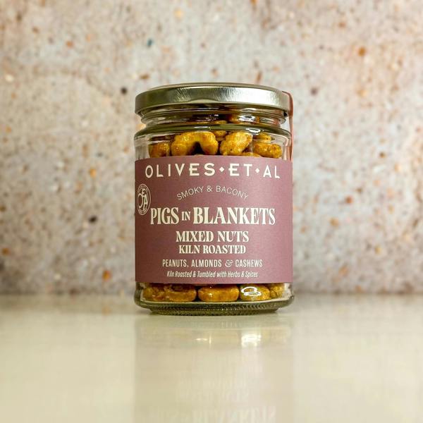 A glass jar of Pigs In Blankets Mixed Nuts sitting on a light counter against a speckled stone backdrop.