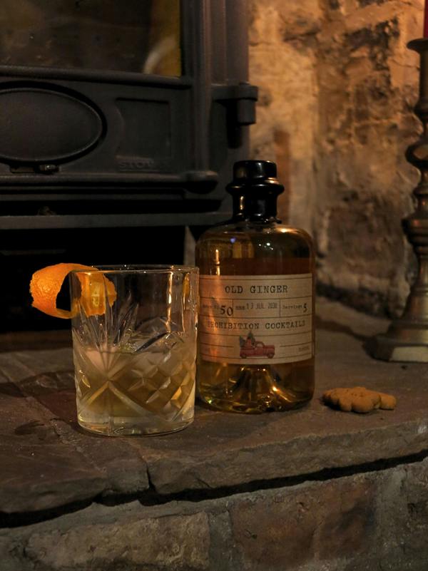 A bottle of Old Ginger Prohibition Cocktails next to a cut glass filled with amber liquid and an orange peel garnish, set on a stone surface in front of a rustic fireplace.