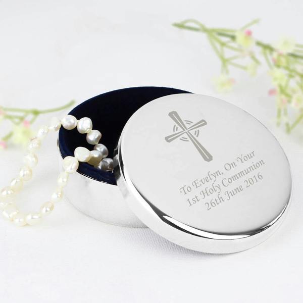 A circular silver trinket box with a cross engraved on the lid, resting on a white surface with small white flowers in the background. The lid is open, revealing the blue flock fabric interior and a string of white pearls.