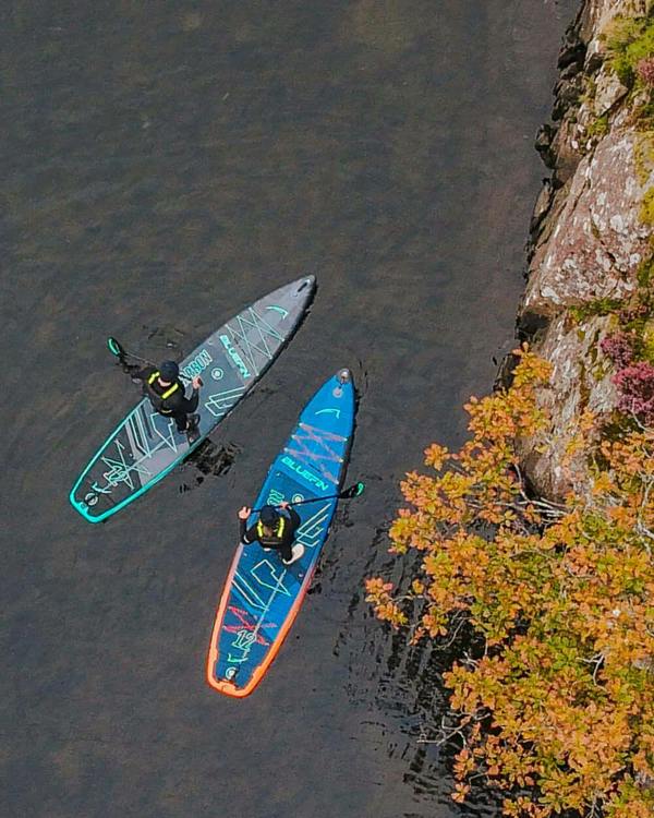 Two paddleboarders on calm water, one in a black and teal board, the other in blue with orange trim.