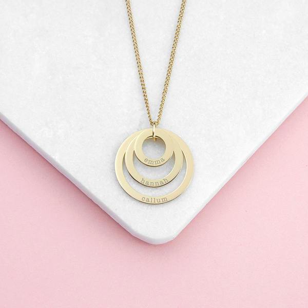 A close-up shot of a three-tier silver nesting family ring pendant necklace engraved with the names 'OLLIE', 'WILLIAM', and 'ZOE' displayed vertically down the face of each concentric loop, resting neatly on a cornered white tile over a soft pink background.
