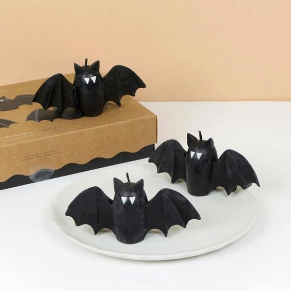 Set of three black bat shaped candles with white fangs sitting on a white dish and kraft box