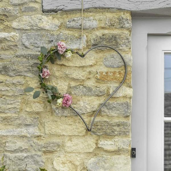 A dark steel heart-shaped frame decorated with pink flowers and eucalyptus leaves, hanging on a rustic stone wall.