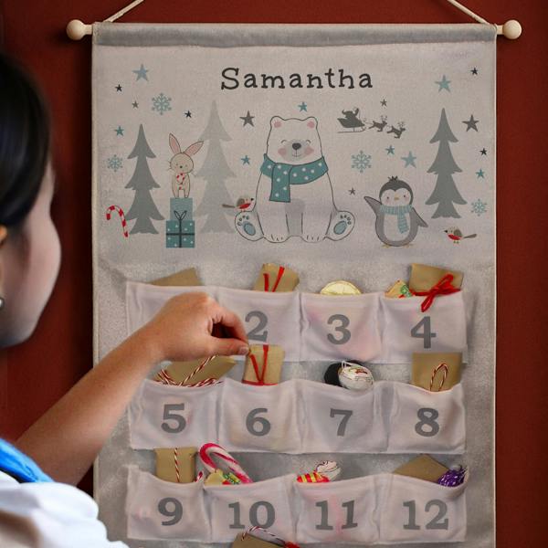 A close-up of a fabric advent calendar hanging on a dark red wall with a hand reaching into pocket number two, featuring a polar bear design at the top printed with the name Samantha.