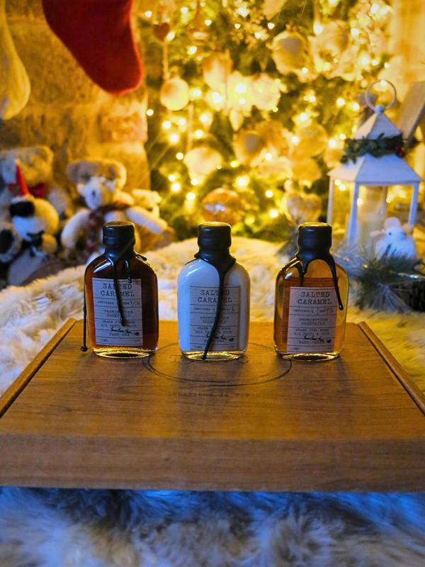 Three small glass bottles with black caps and cream labels reading 'Salted Caramel' are arranged on a wooden tray in a cozy holiday setting with a lit Christmas tree, teddy bears, and warm ambient lights in the background.