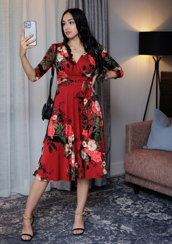 This is a young lady wearing a Mock wrap midi dress in a Red floral design, with 3/4 length sleeves, a gathered shoulder and under bust area, with a tie, secured in a bow. It has a V neck front. The dress has a midi length full skirt in a retro style. The model is wearing black strappy heels, with a small black shoulder bag. She has long dark hair and is taking a picture, she is silhouetted 