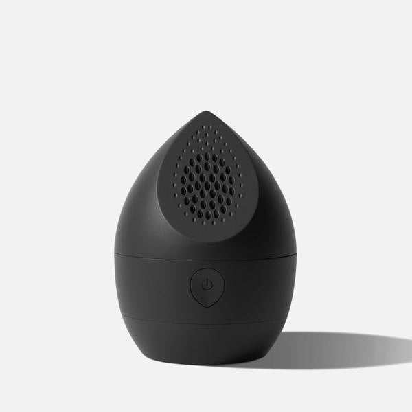 A compact, black, teardrop-shaped Fridja f40 Fabric Shaver from the Garment Care Trio rests on a light surface. Its power button sits on the front, with a perforated speaker-like grille on top, echoing a fabric shaver’s classic design.