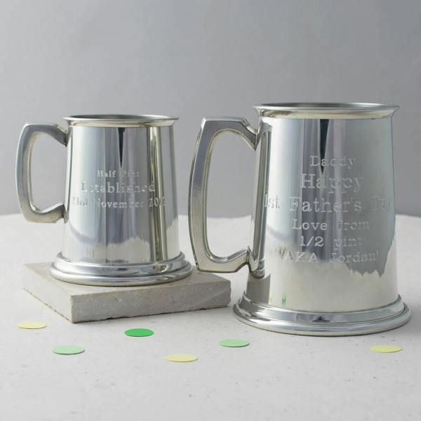 Two polished pewter tankards in half-pint and full-pint sizes, featuring custom engravings for an anniversary and a first Father's Day, sitting on a stone plinth.